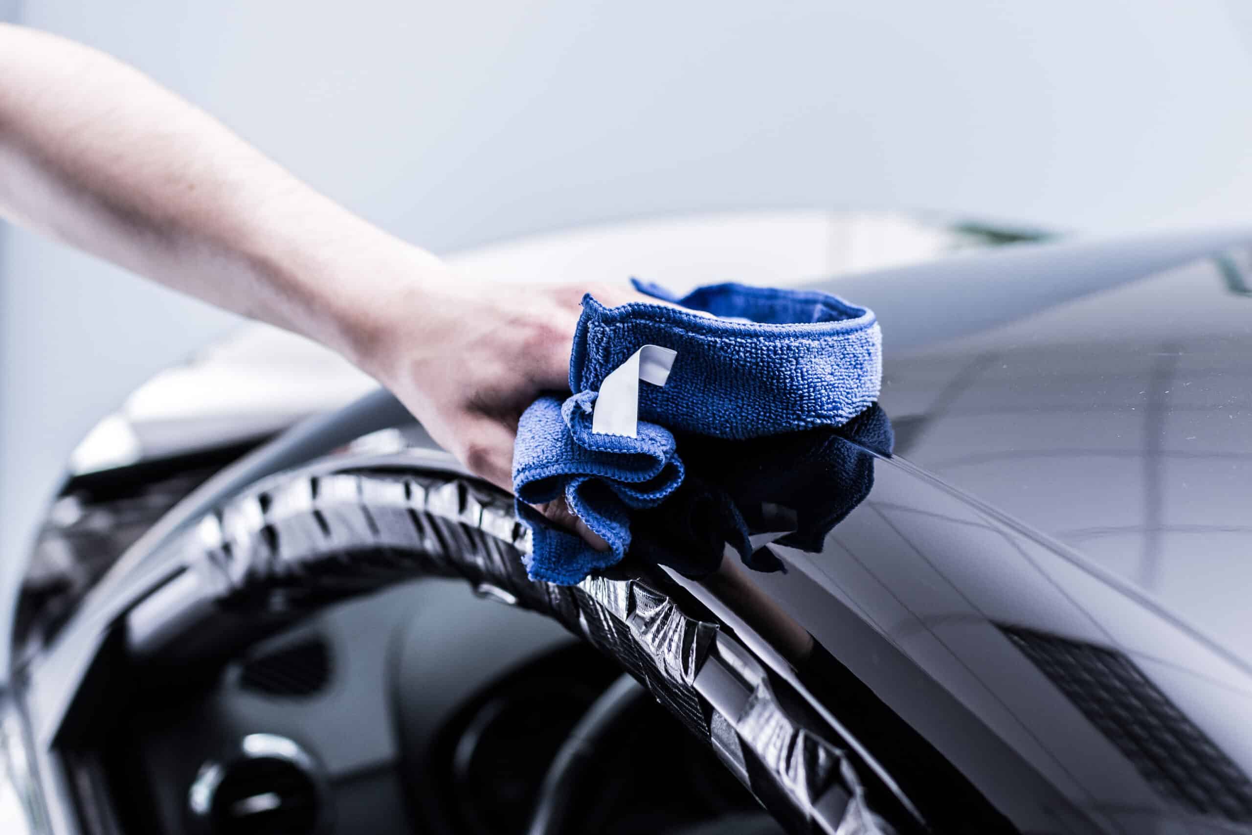 Main essuyant une carrosserie noire très brillante avec un chiffon microfibre bleu lors d'une préparation ou d'un detailing automobile.