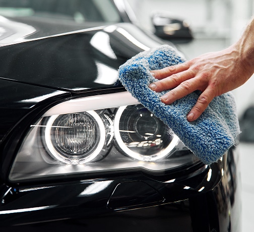 Main polissant un phare de voiture noir brillant avec un chiffon microfibre bleu texturé pour un entretien automobile.